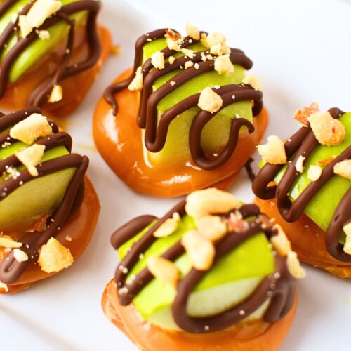 Hand holding a single caramel apple pretzel bite with chocolate drizzle and peanuts with platter of bites in the background