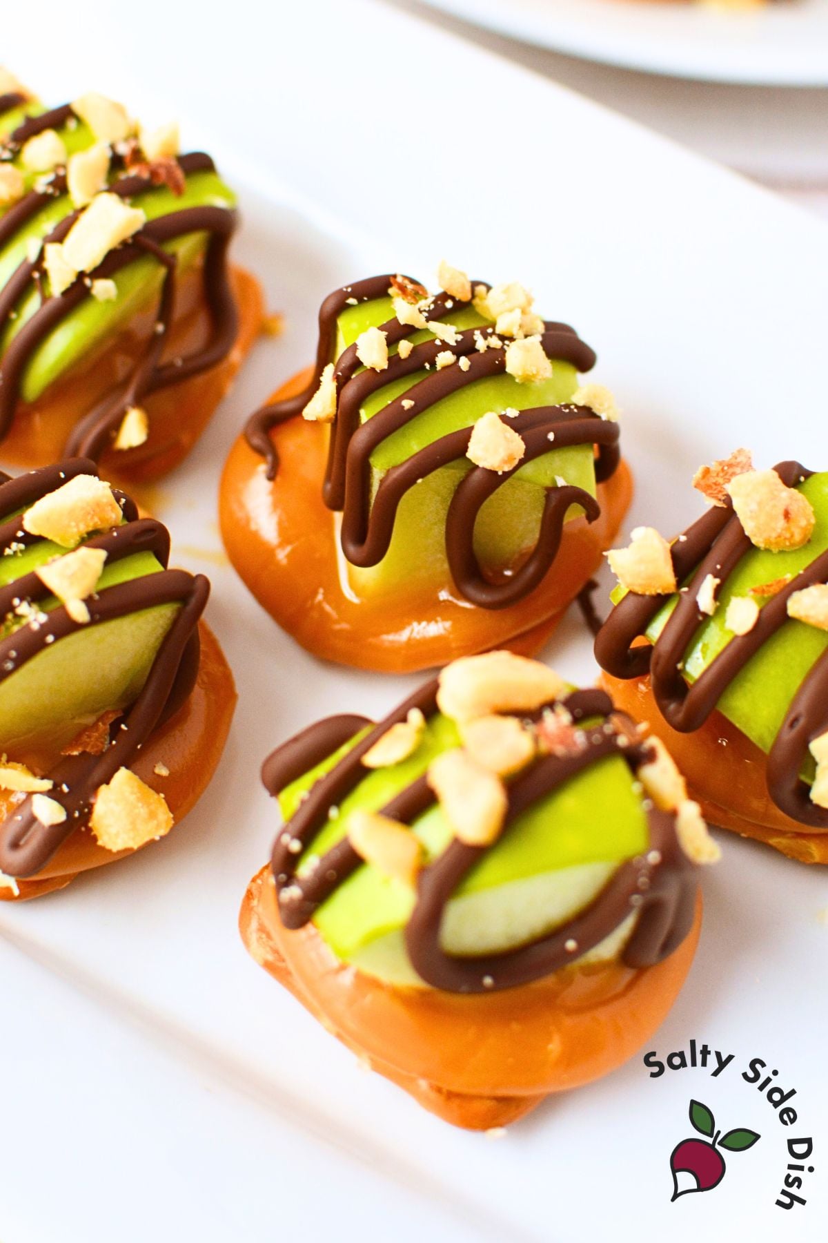 Hand holding a single caramel apple pretzel bite with chocolate drizzle and peanuts with platter of bites in the background