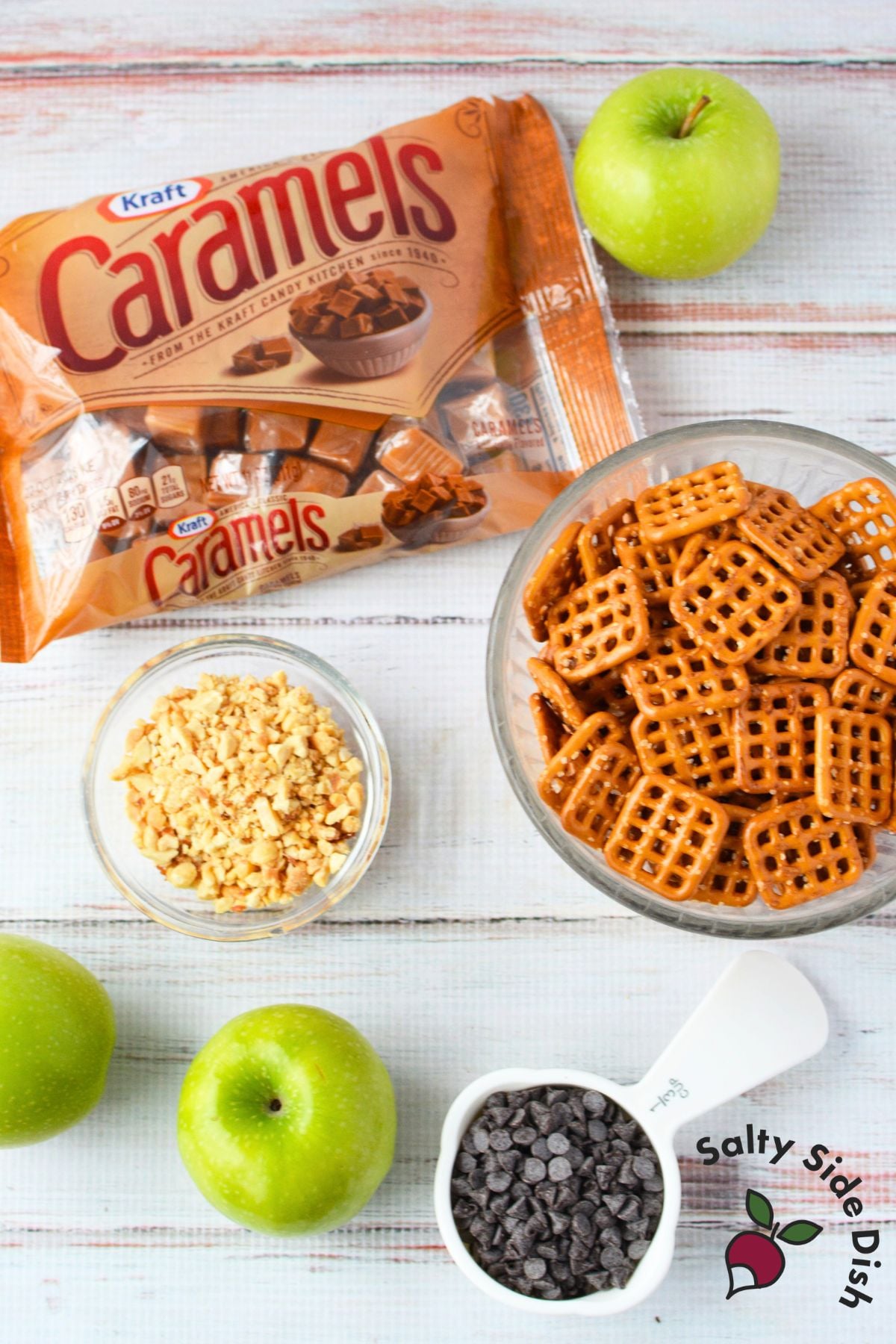 Kraft caramels bag, square pretzel snaps, granny smith apples, mini chocolate chips, and chopped peanuts arranged on a white wood surface