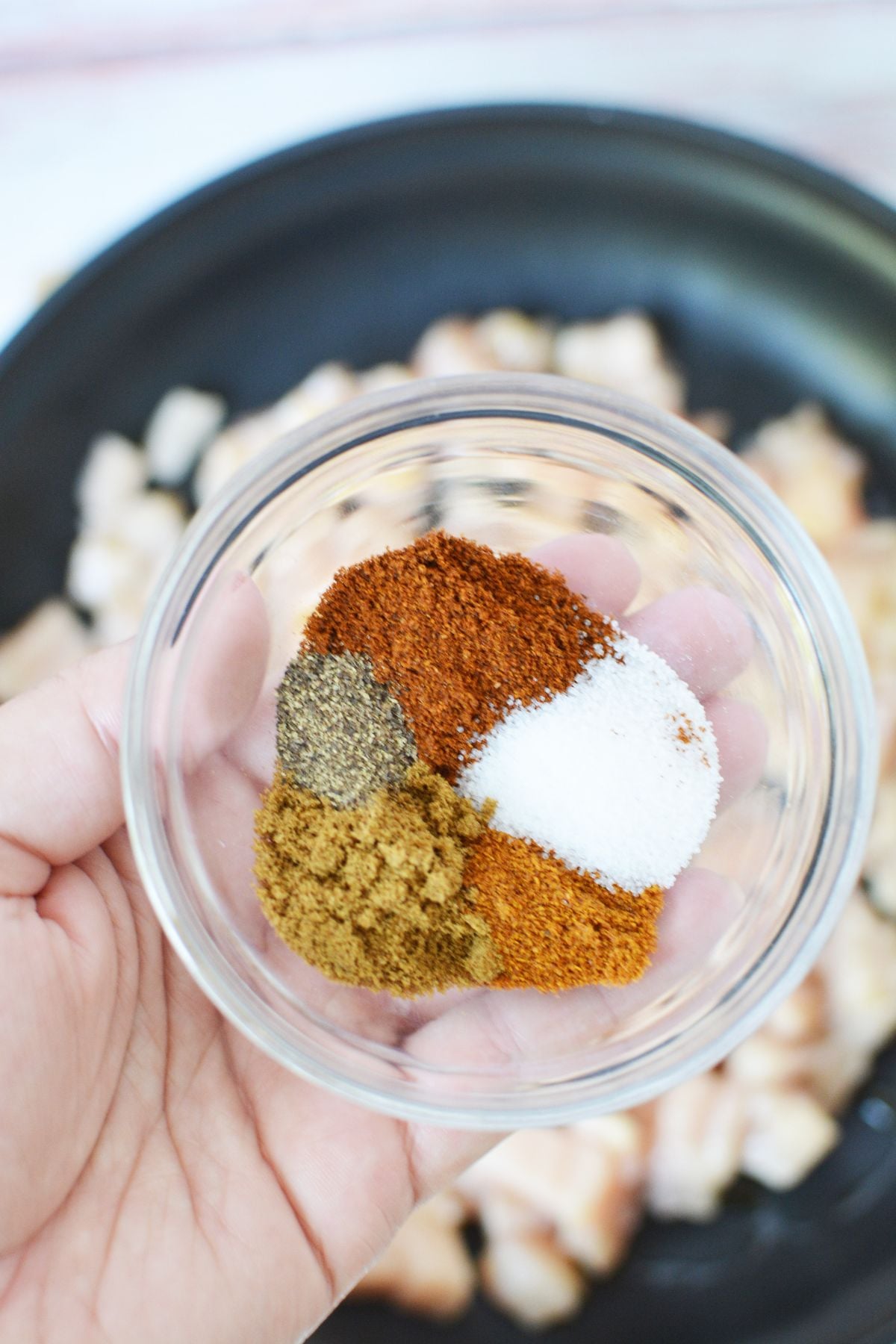 Taco seasoning spices including chili powder, cumin, cayenne, salt and pepper in a glass bowl with raw chicken in background
