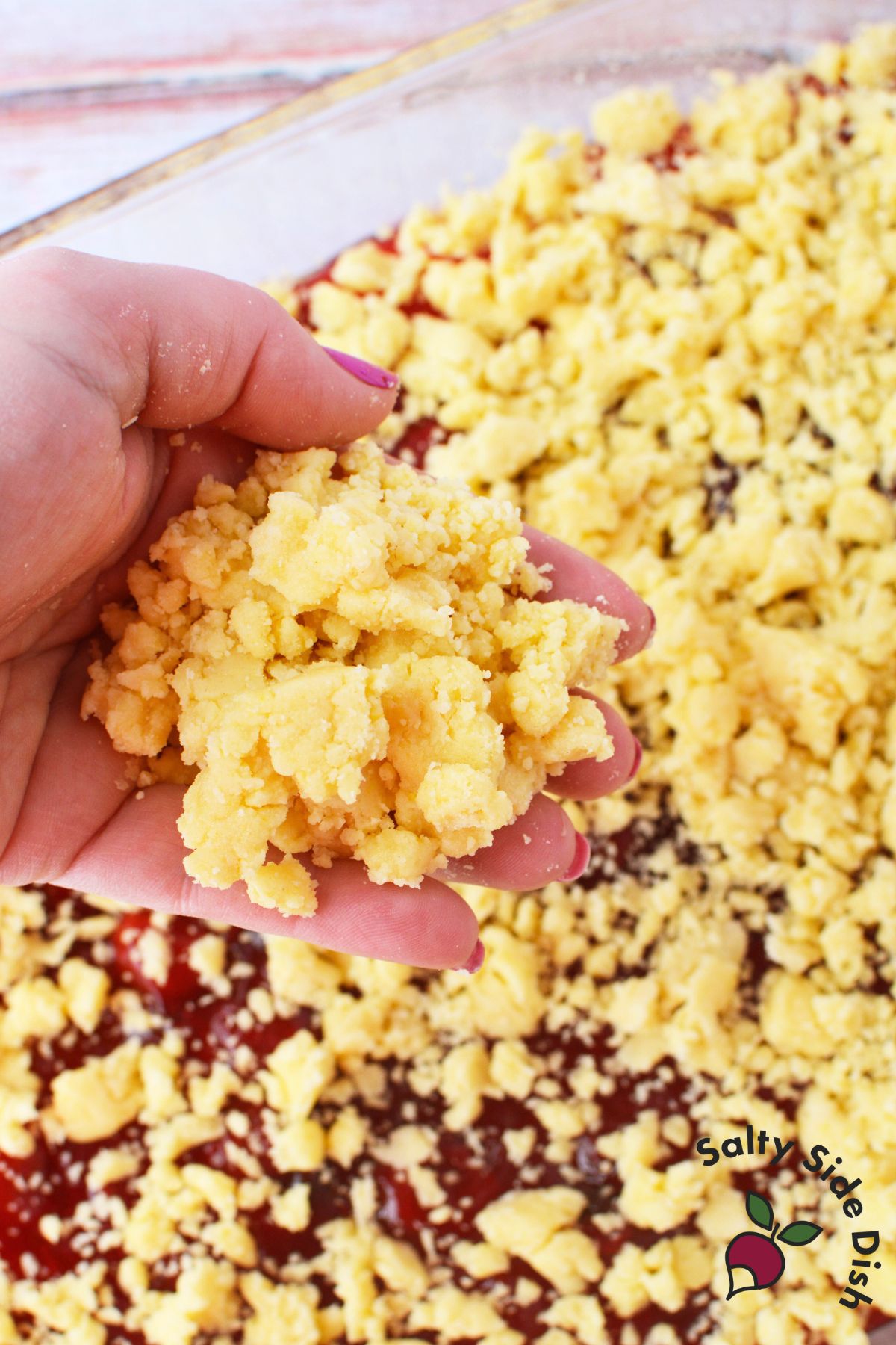 Hand holding a clump of lemon cake mix and butter crumb topping over a 9x13 baking dish of strawberry lemon dump cake before baking