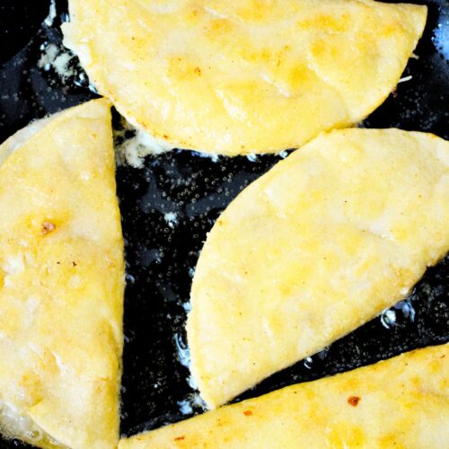 Three folded chicken tacos frying in vegetable oil in black skillet until golden and crispy