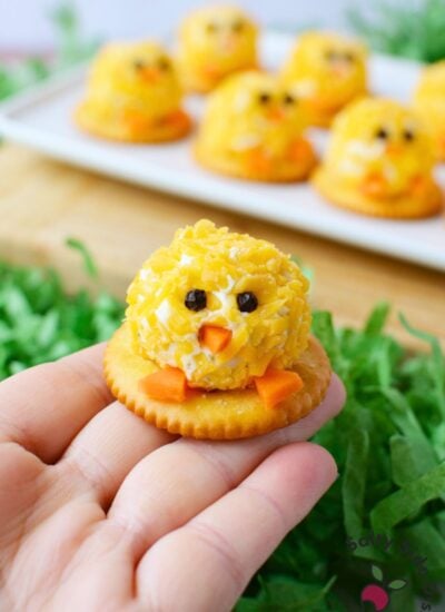 hand holding a single mini chick cheeseball bite on a round cracker with peppercorn eyes carrot beak and carrot feet with a tray of Easter cheeseball bites in the background