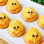 mini chick cheeseball bites on a white serving tray decorated with peppercorn eyes and carrot beaks surrounded by green Easter grass