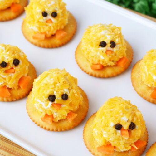 mini chick cheeseball bites on a white serving tray decorated with peppercorn eyes and carrot beaks surrounded by green Easter grass