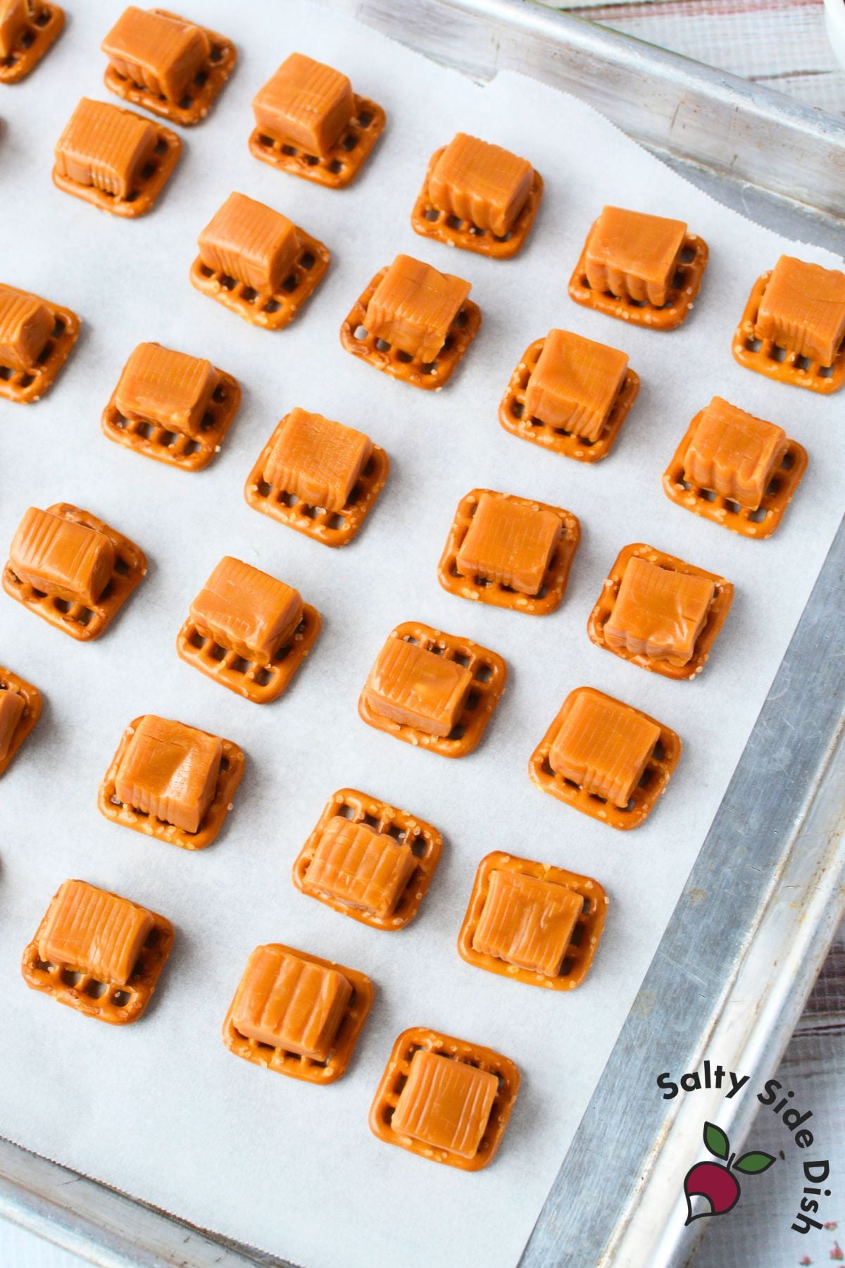 Square pretzel snaps topped with unwrapped caramel squares arranged in rows on a parchment lined baking sheet