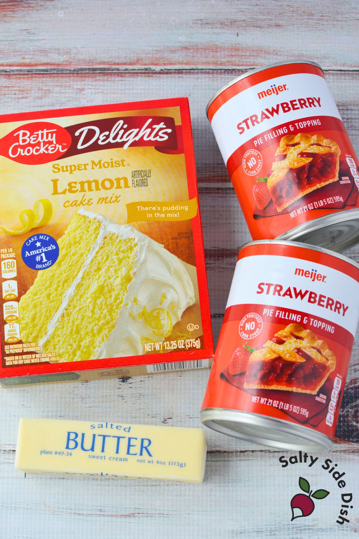 Three ingredients for strawberry lemon dump cake laid out on a white wood surface - Betty Crocker lemon cake mix, two cans of Meijer strawberry pie filling, and a stick of salted butter