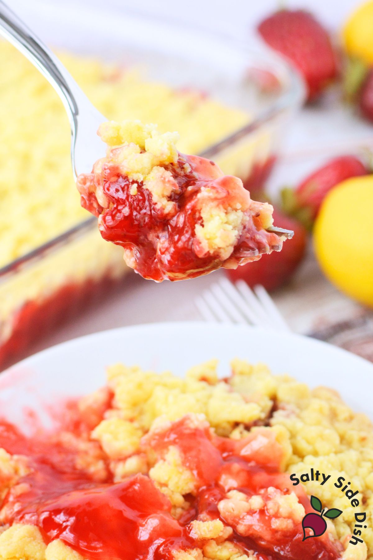 Fork holding a bite of baked strawberry lemon dump cake with gooey strawberry filling and golden lemon crumb topping, with fresh strawberries and a lemon in the background