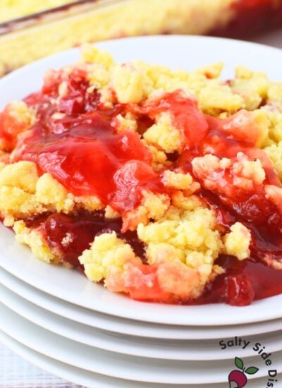 Serving of strawberry lemon dump cake on a white plate stacked on other plates, showing bubbly strawberry filling bubbling through golden lemon cake mix topping, with the baking dish in the background