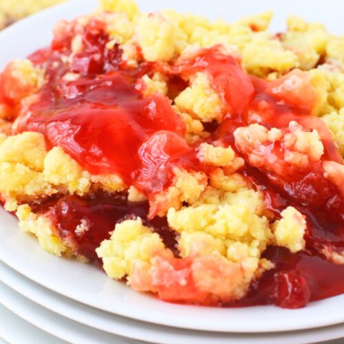 Serving of strawberry lemon dump cake on a white plate stacked on other plates, showing bubbly strawberry filling bubbling through golden lemon cake mix topping, with the baking dish in the background