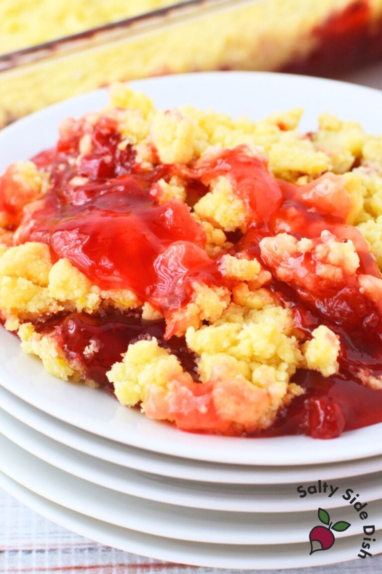 Serving of strawberry lemon dump cake on a white plate stacked on other plates, showing bubbly strawberry filling bubbling through golden lemon cake mix topping, with the baking dish in the background