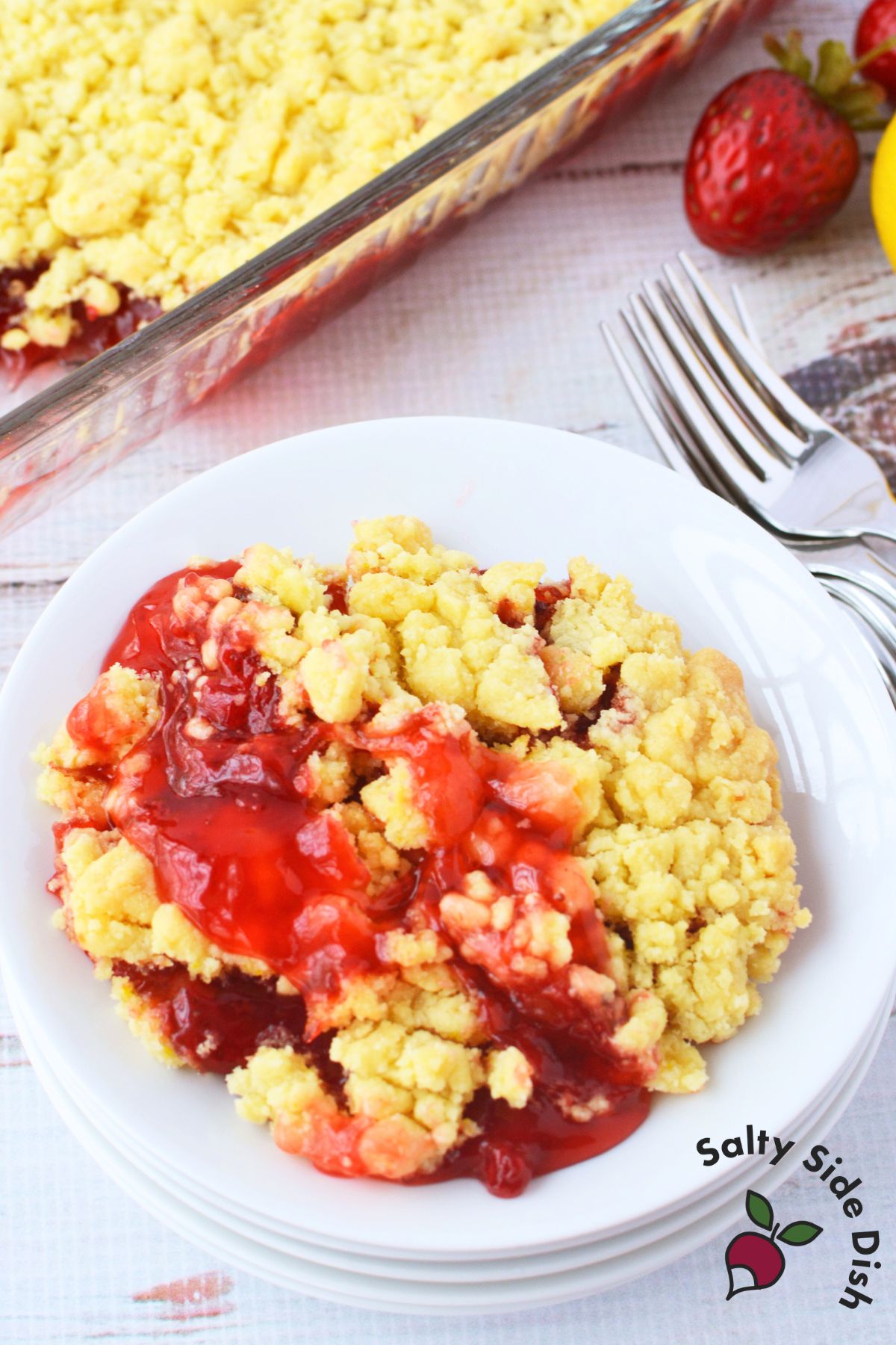 Overhead view of a serving of strawberry lemon dump cake on a white plate with golden lemon crumb topping and bright red strawberry filling, fork and fresh strawberries alongside the 9x13 baking dish