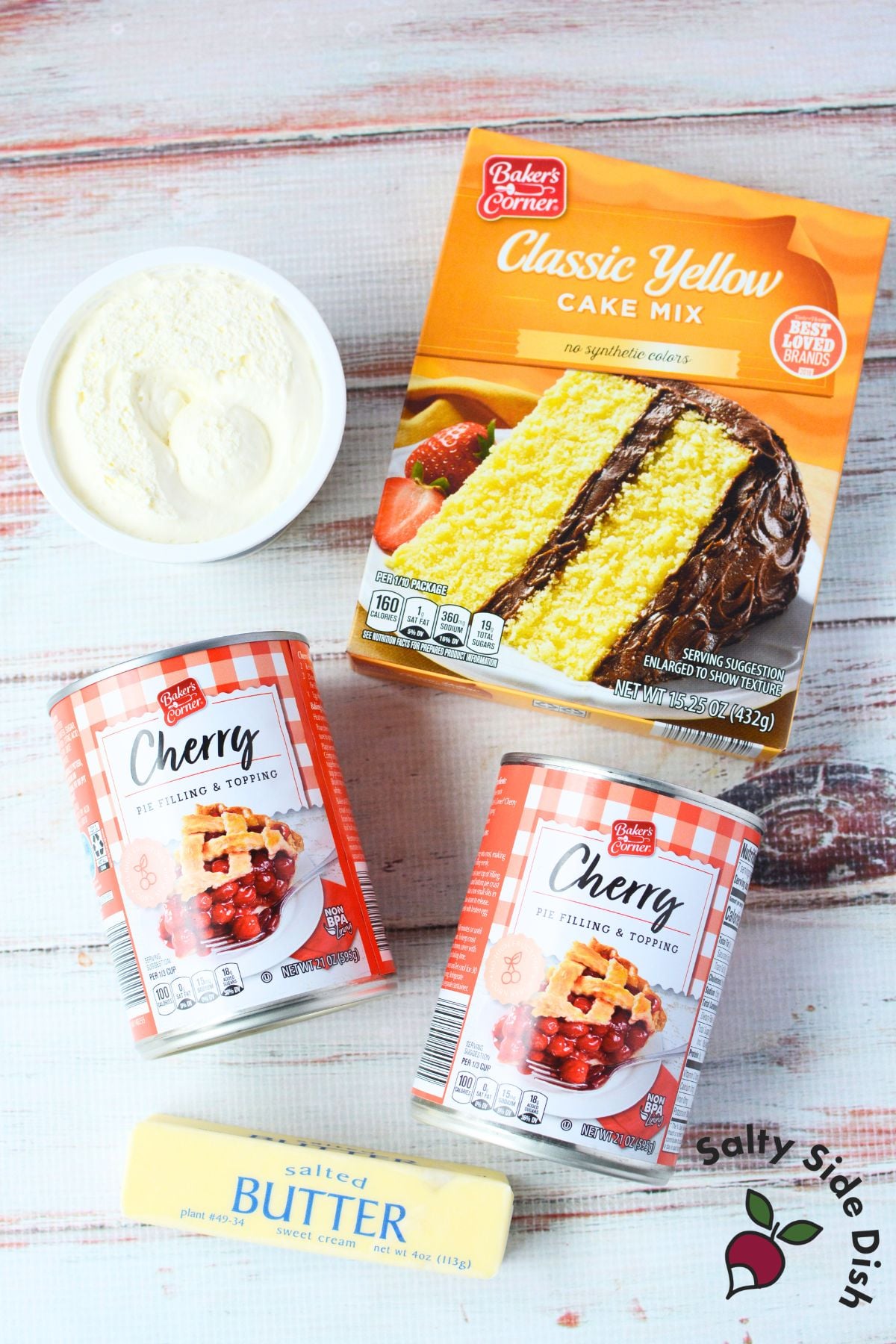 ingredients for cherry cheesecake dump cake including yellow cake mix, two cans of cherry pie filling, cream cheese, and salted butter on a white wood surface