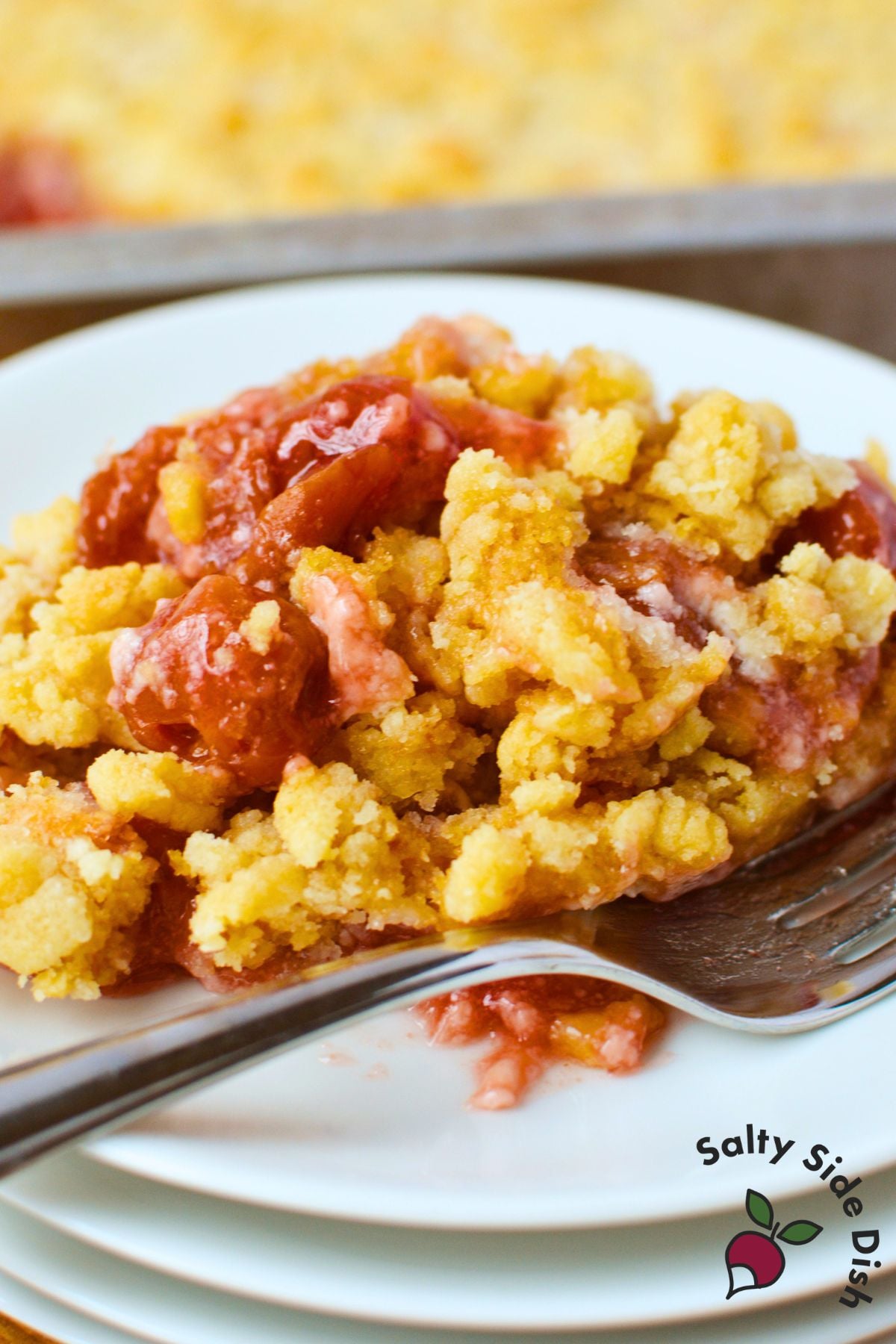 plated serving of cherry cheesecake dump cake with buttery golden crumble and cherry filling, fork resting on white plate