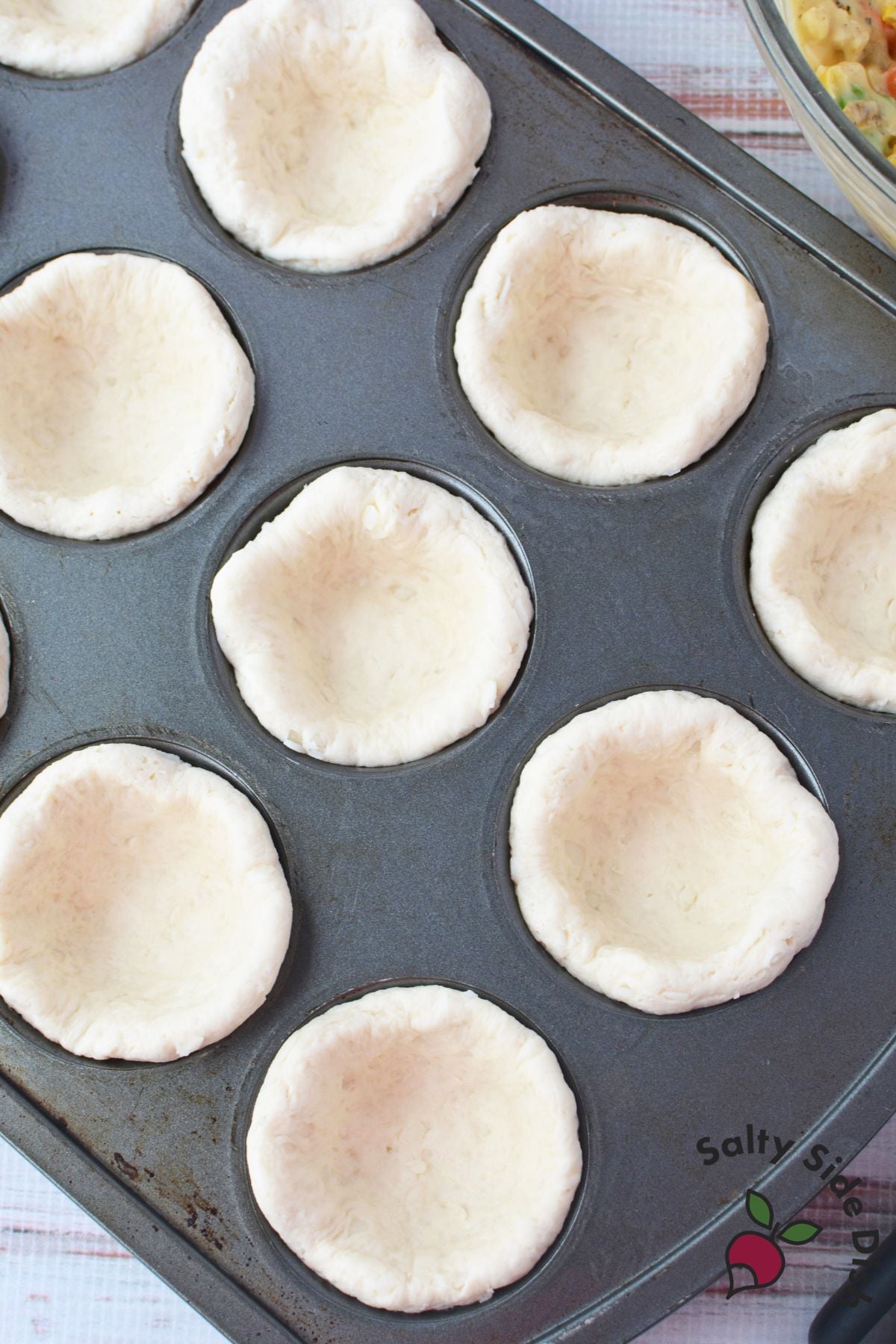 Jumbo buttermilk biscuits pressed into a greased 12-cup muffin tin forming cup shapes ready for filling