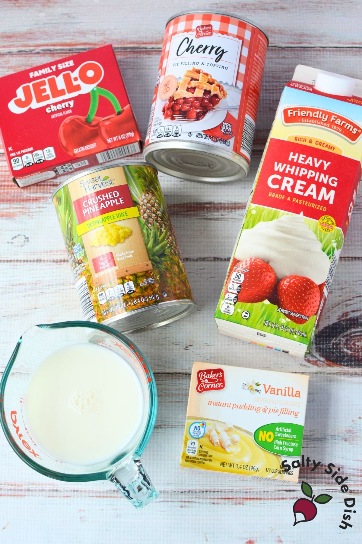 Ingredients for cherry jello salad laid out on a white wood surface including cherry Jell-O, cherry pie filling, crushed pineapple, vanilla instant pudding, heavy whipping cream, and milk