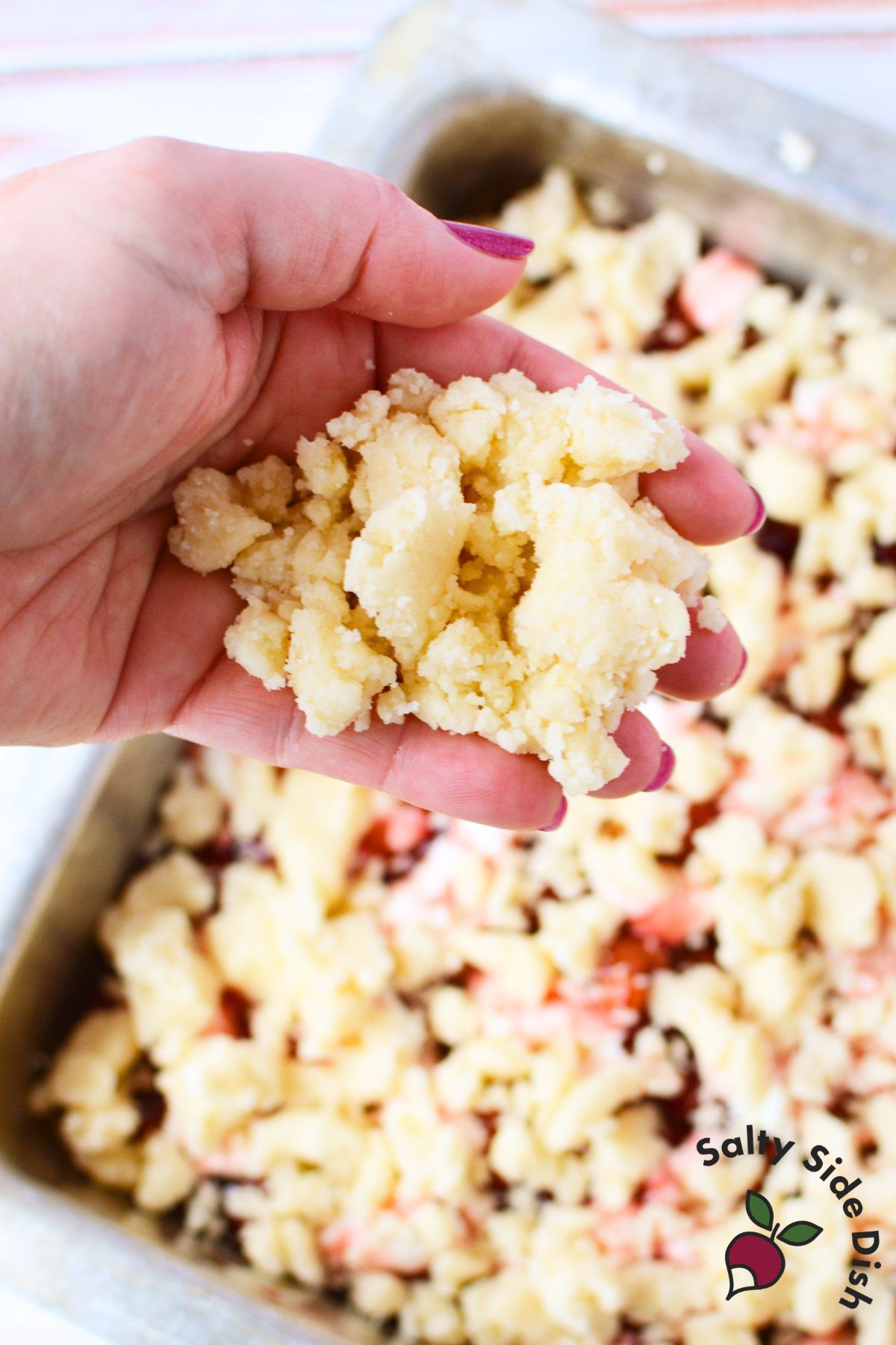 hand holding yellow cake mix crumble topping over baking pan filled with cherry and cream cheese layers