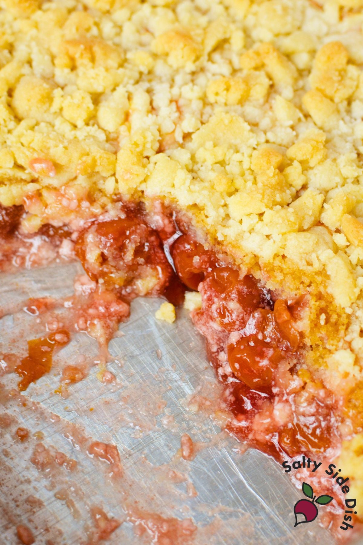 close up of cherry cheesecake dump cake in baking pan with golden crumble topping and section removed showing cherry filling layers
