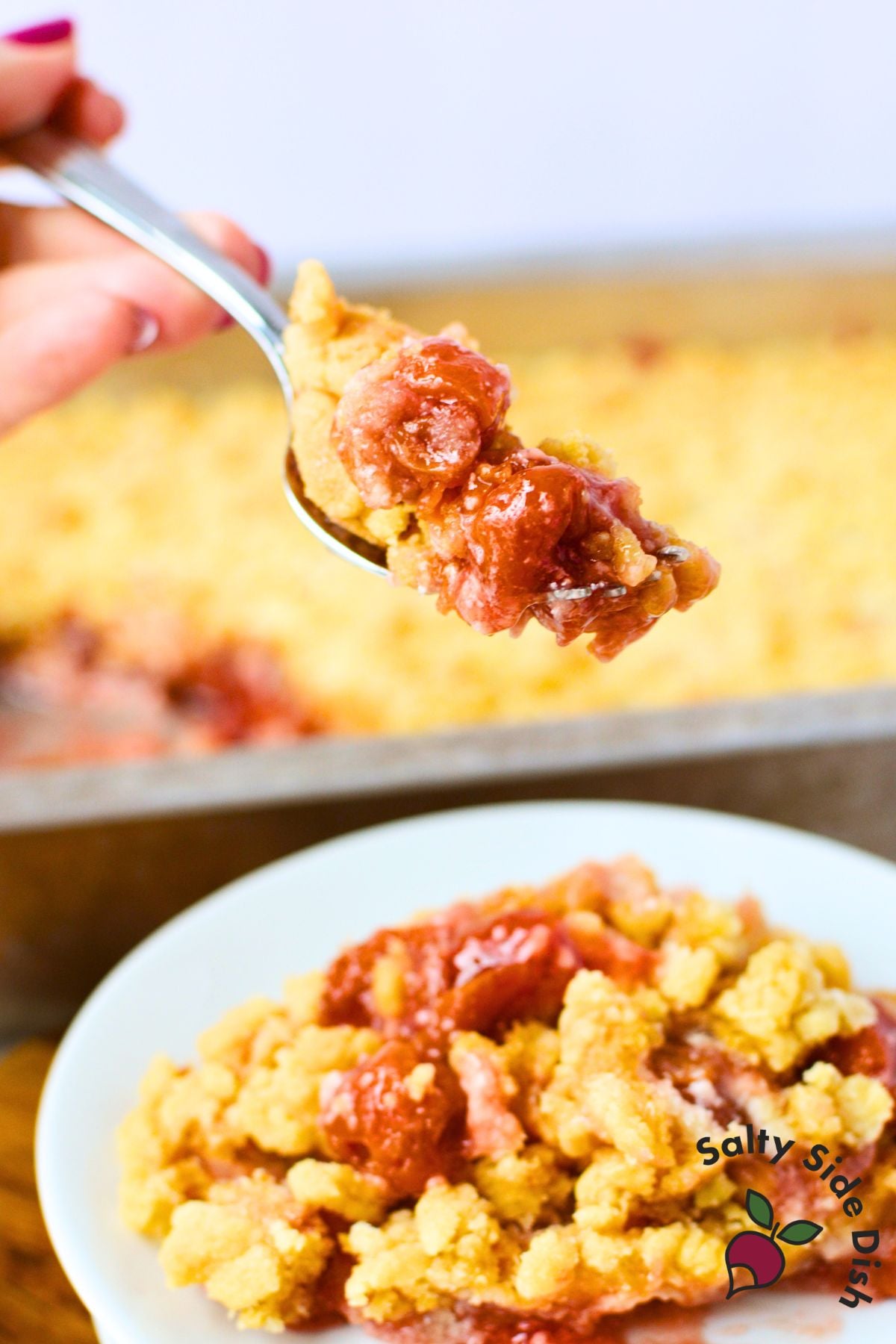 spoonful of cherry cheesecake dump cake held over a white plate with baking pan of golden crumble topping in background