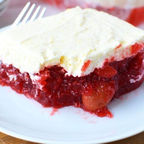 Slice of cherry pineapple jello salad on a white plate with a fork, showing a whole cherry visible in the deep red jello layer beneath vanilla pudding topping