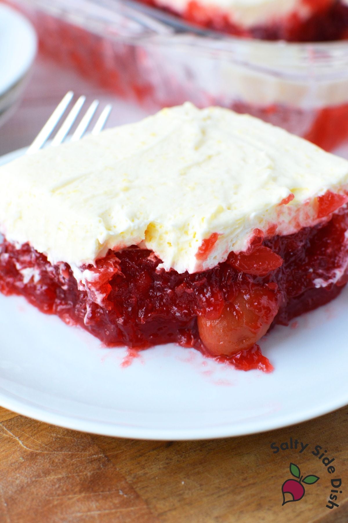 Slice of cherry pineapple jello salad on a white plate with a fork, showing a whole cherry visible in the deep red jello layer beneath vanilla pudding topping