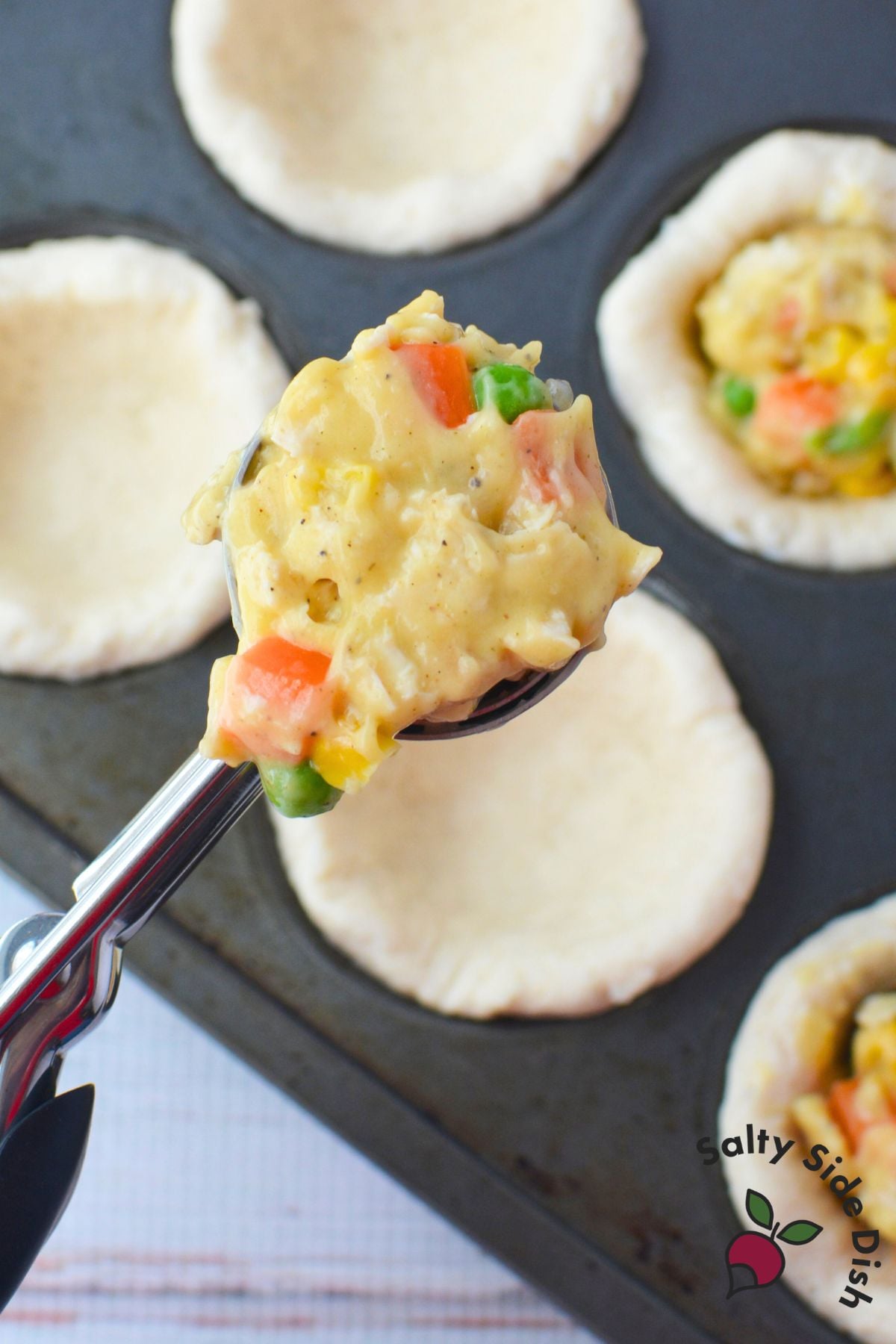 Scooping creamy chicken and vegetable filling into unbaked biscuit cups in a muffin tin