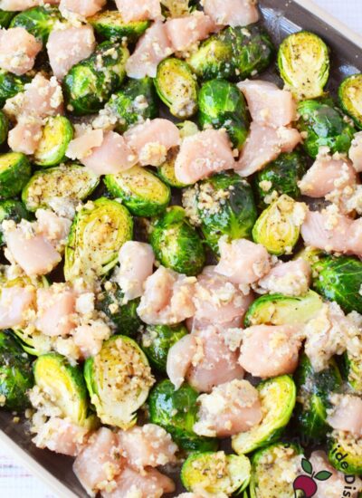 raw chicken and brussels sprouts seasoned with garlic butter spread out on a sheet pan ready to bake
