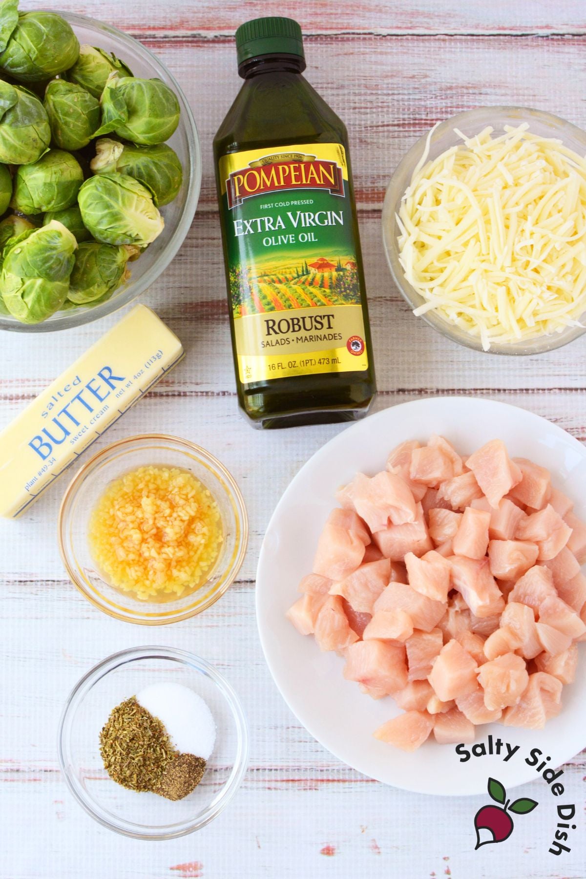 ingredients for sheet pan chicken and brussels sprouts including raw chicken breast, fresh brussels sprouts, butter, olive oil, minced garlic, shredded gruyere cheese, and seasonings