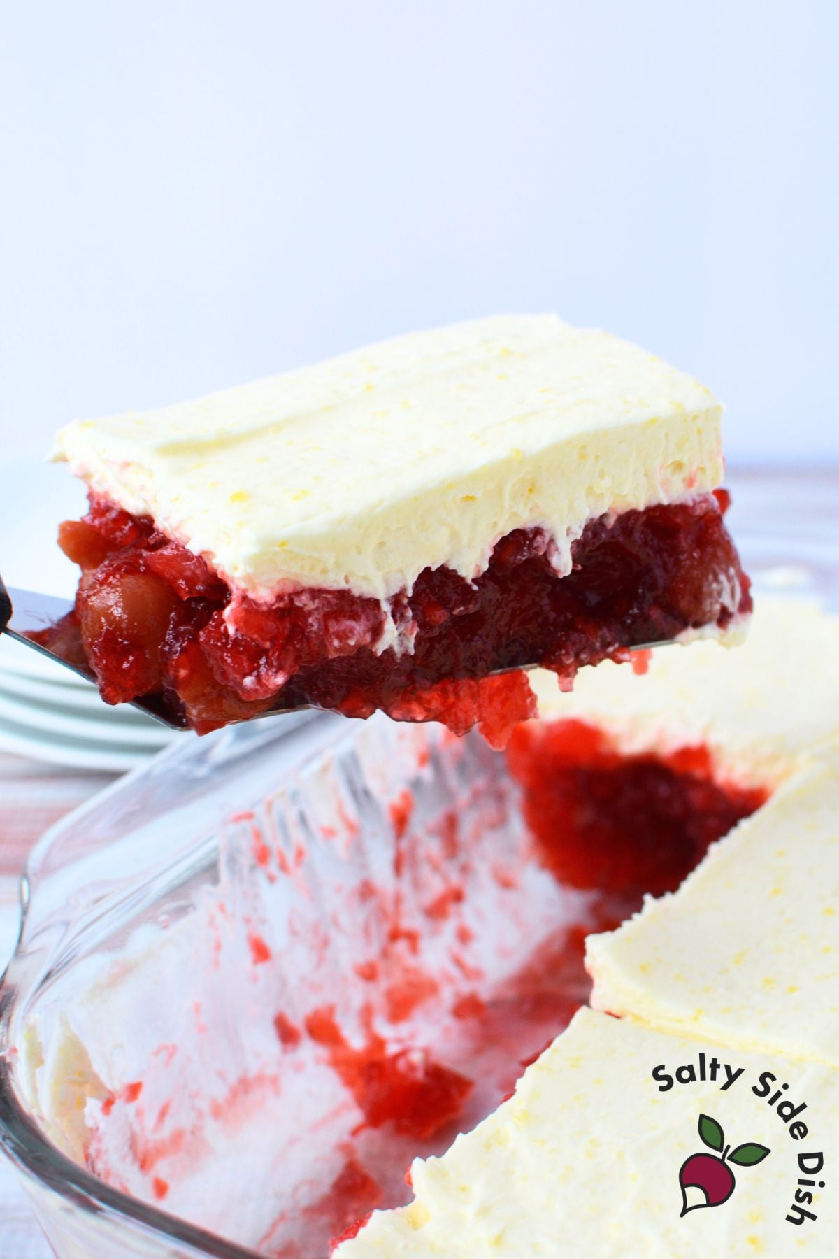Spatula lifting a square serving of cherry pineapple jello salad from a glass baking dish, showing the two distinct layers of red cherry jello and creamy vanilla topping