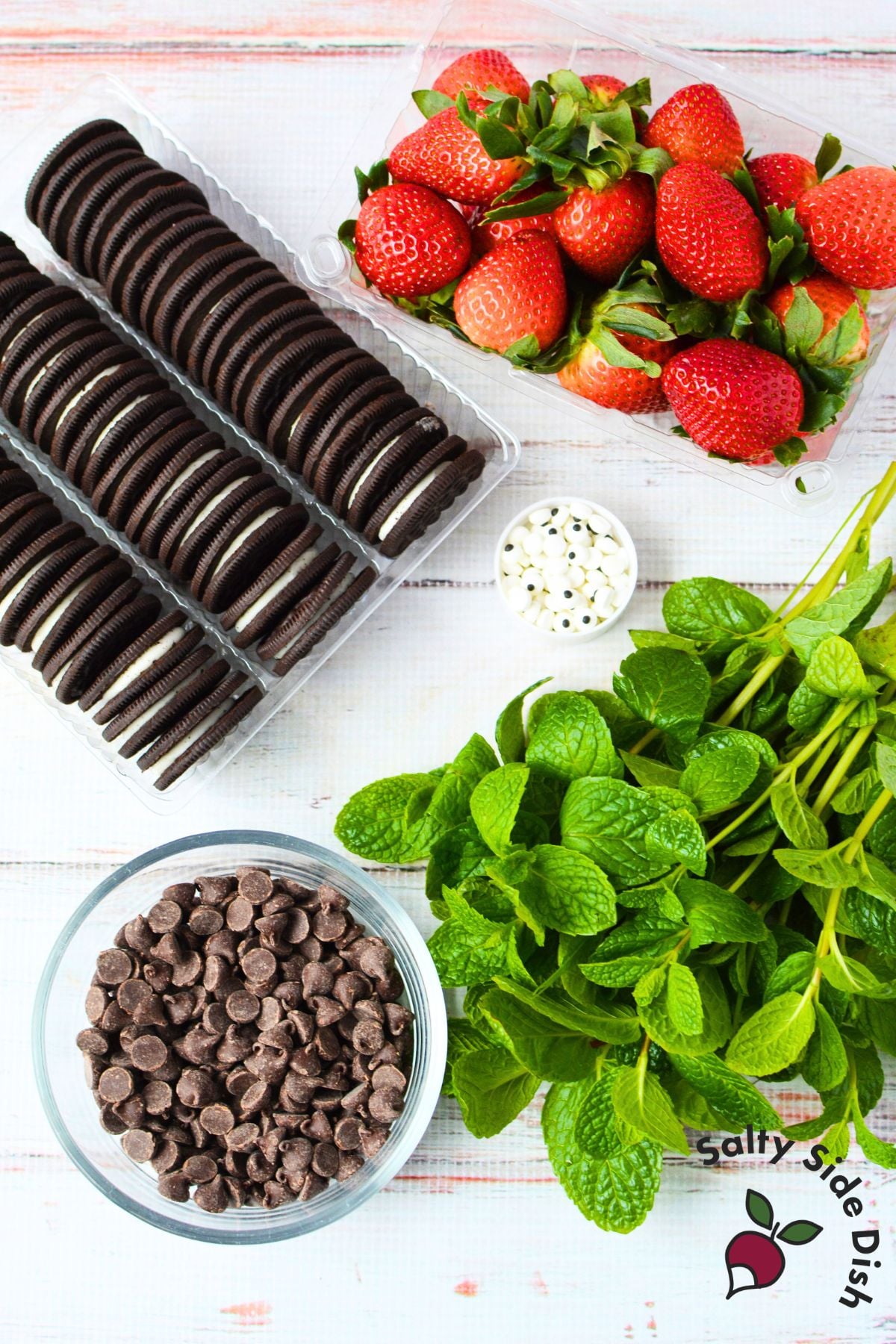 Ingredients for strawberry ladybugs including fresh strawberries, Oreo cookies, chocolate chips, candy eyes, and fresh mint leaves
