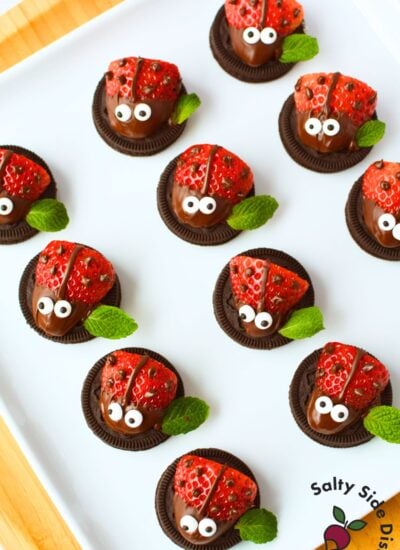 Overhead view of finished strawberry ladybugs on a white platter with chocolate spots, candy eyes, and fresh mint leaf wings