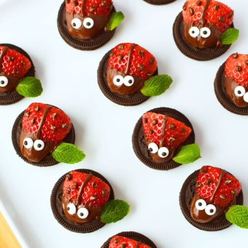 Overhead view of finished strawberry ladybugs on a white platter with chocolate spots, candy eyes, and fresh mint leaf wings