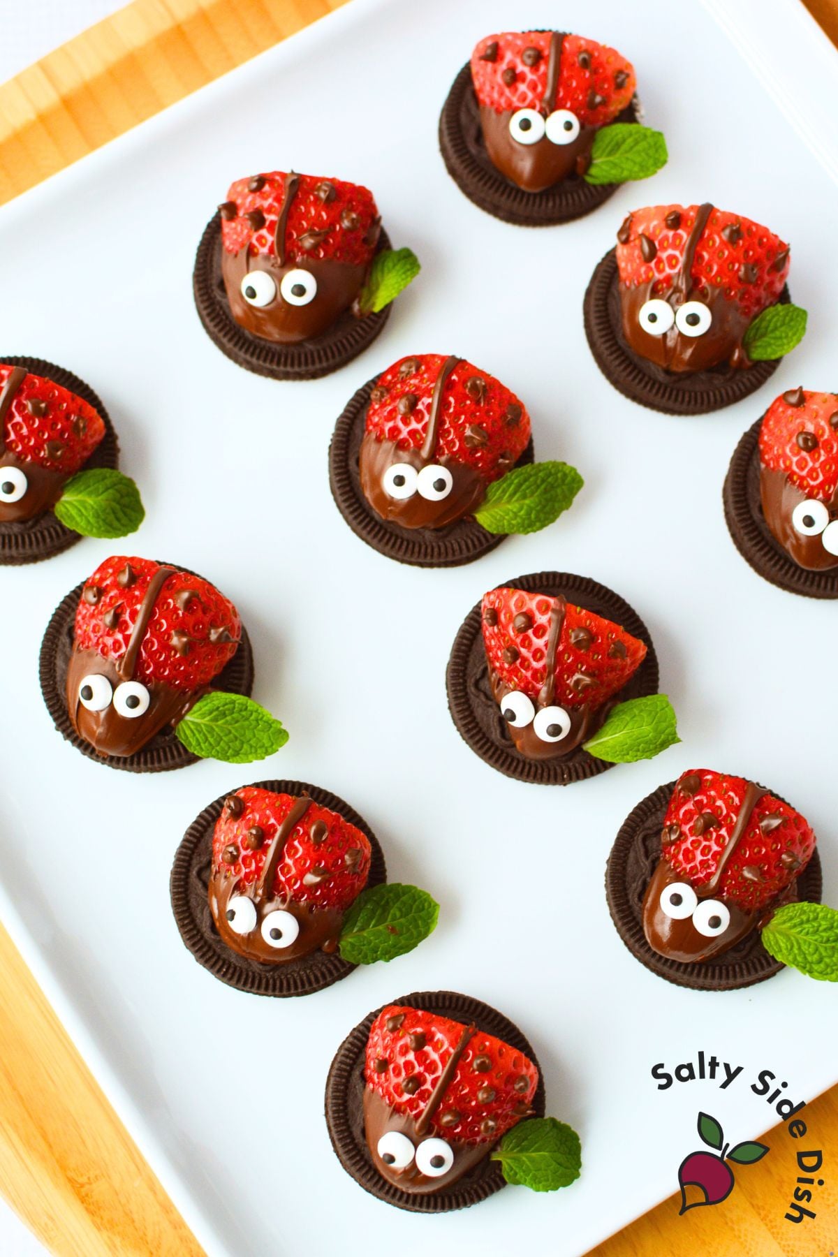 Overhead view of finished strawberry ladybugs on a white platter with chocolate spots, candy eyes, and fresh mint leaf wings