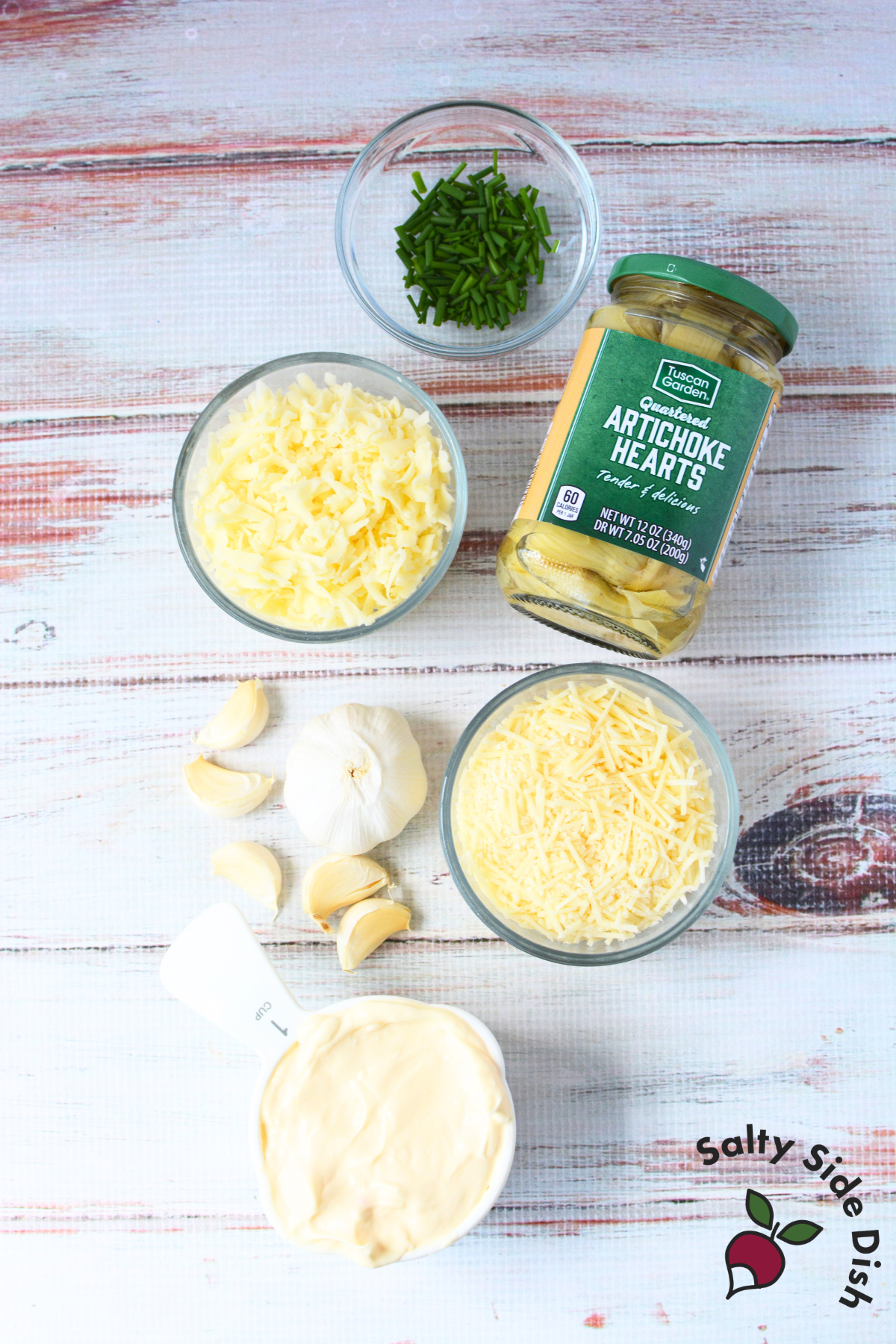 flat lay of artichoke dip ingredients including a jar of quartered artichoke hearts, shredded mozzarella, shredded parmesan, garlic cloves, mayonnaise, and fresh chives