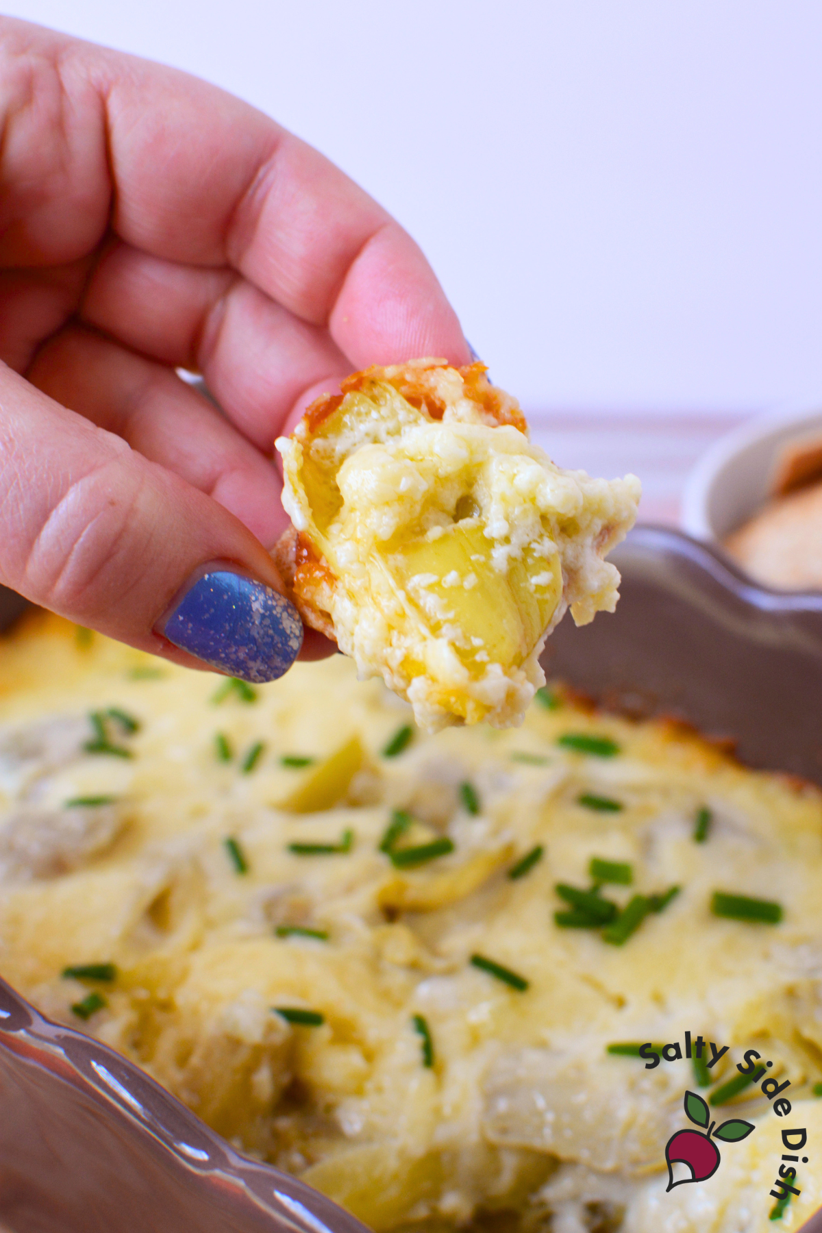 hand holding a cracker loaded with warm baked artichoke dip over a casserole dish