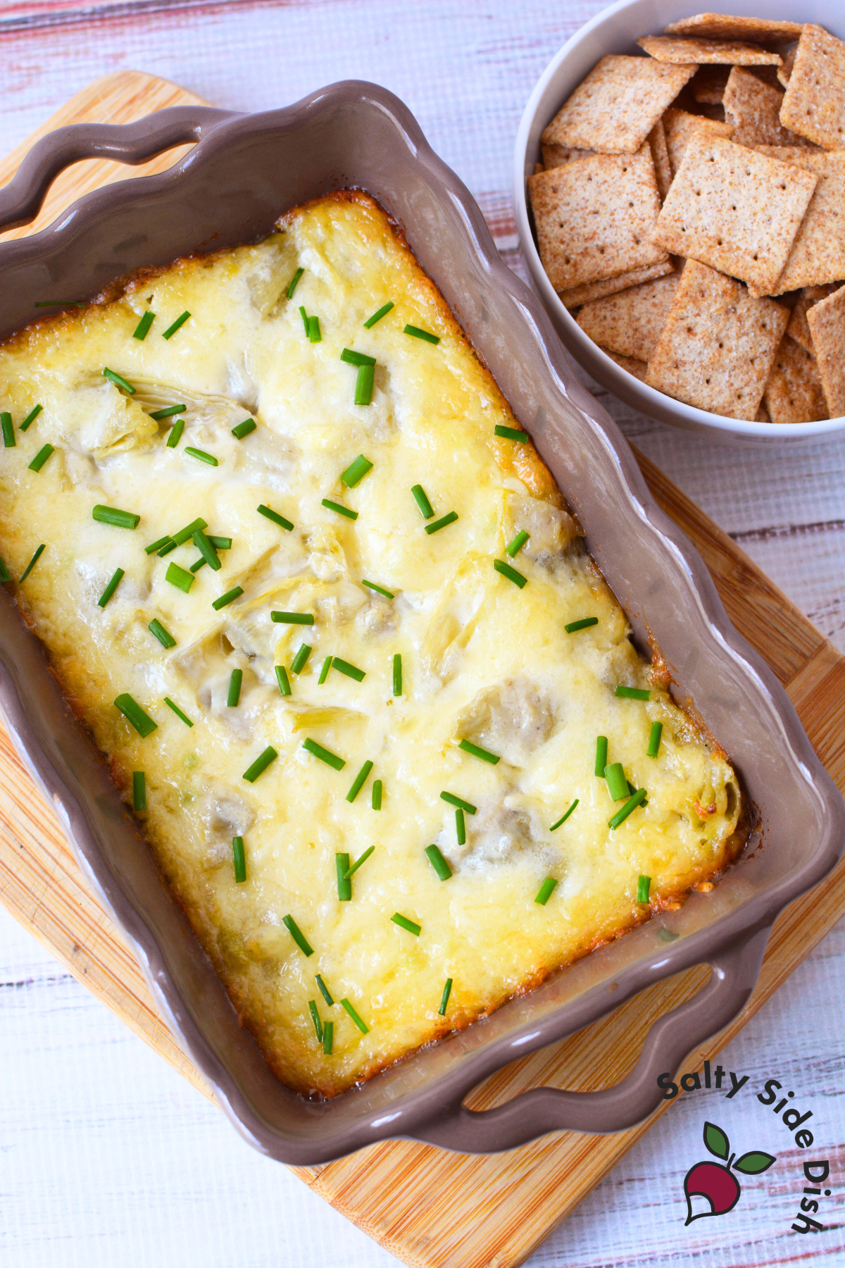 aked artichoke dip with golden bubbly top and fresh chives in a casserole dish served with wheat crackers