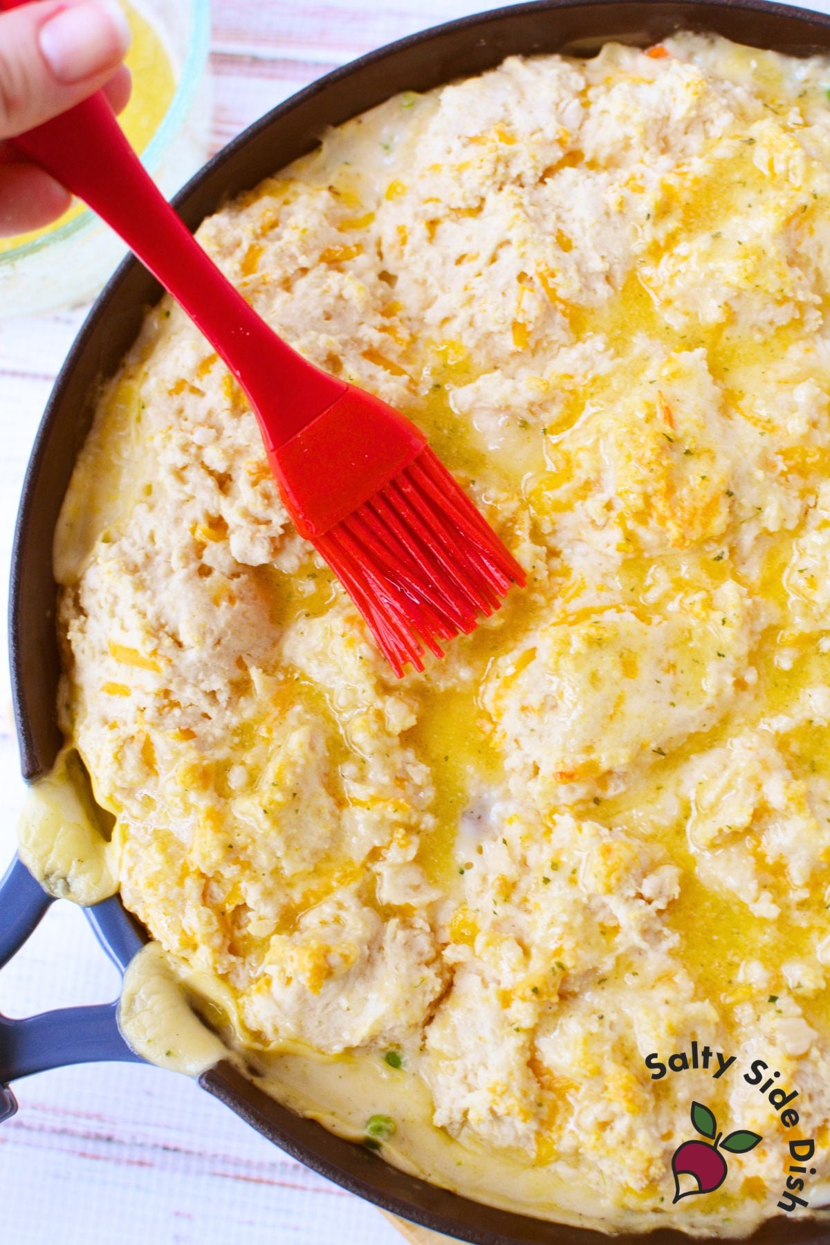 Red silicone pastry brush spreading melted butter over raw Cheddar Bay Biscuit topping on chicken pot pie in a cast iron skillet