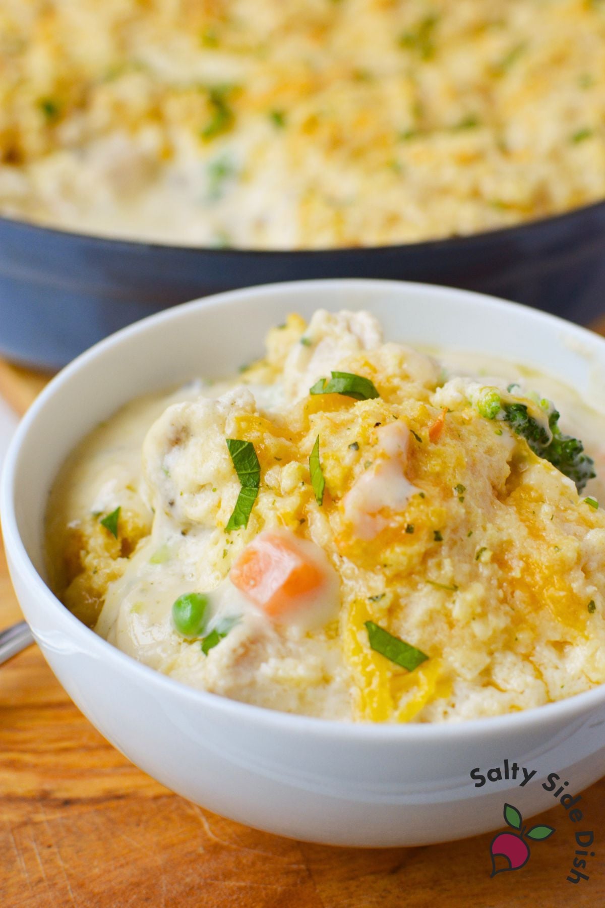 Close up of a white bowl filled with creamy chicken pot pie topped with golden Cheddar Bay Biscuit pieces, shredded cheddar, and fresh parsley with the skillet in the background