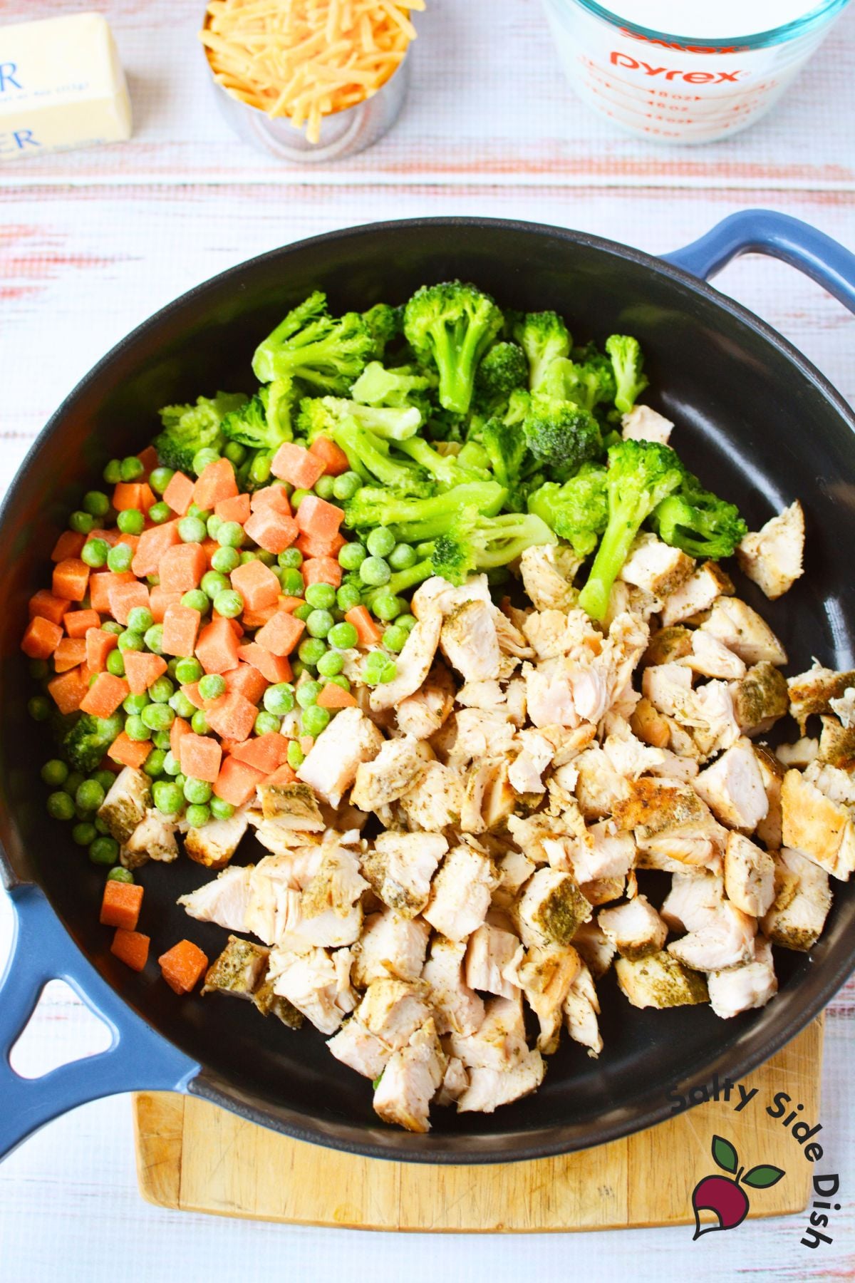 Large cast iron skillet filled with cubed cooked chicken, broccoli florets, and peas and carrots before mixing