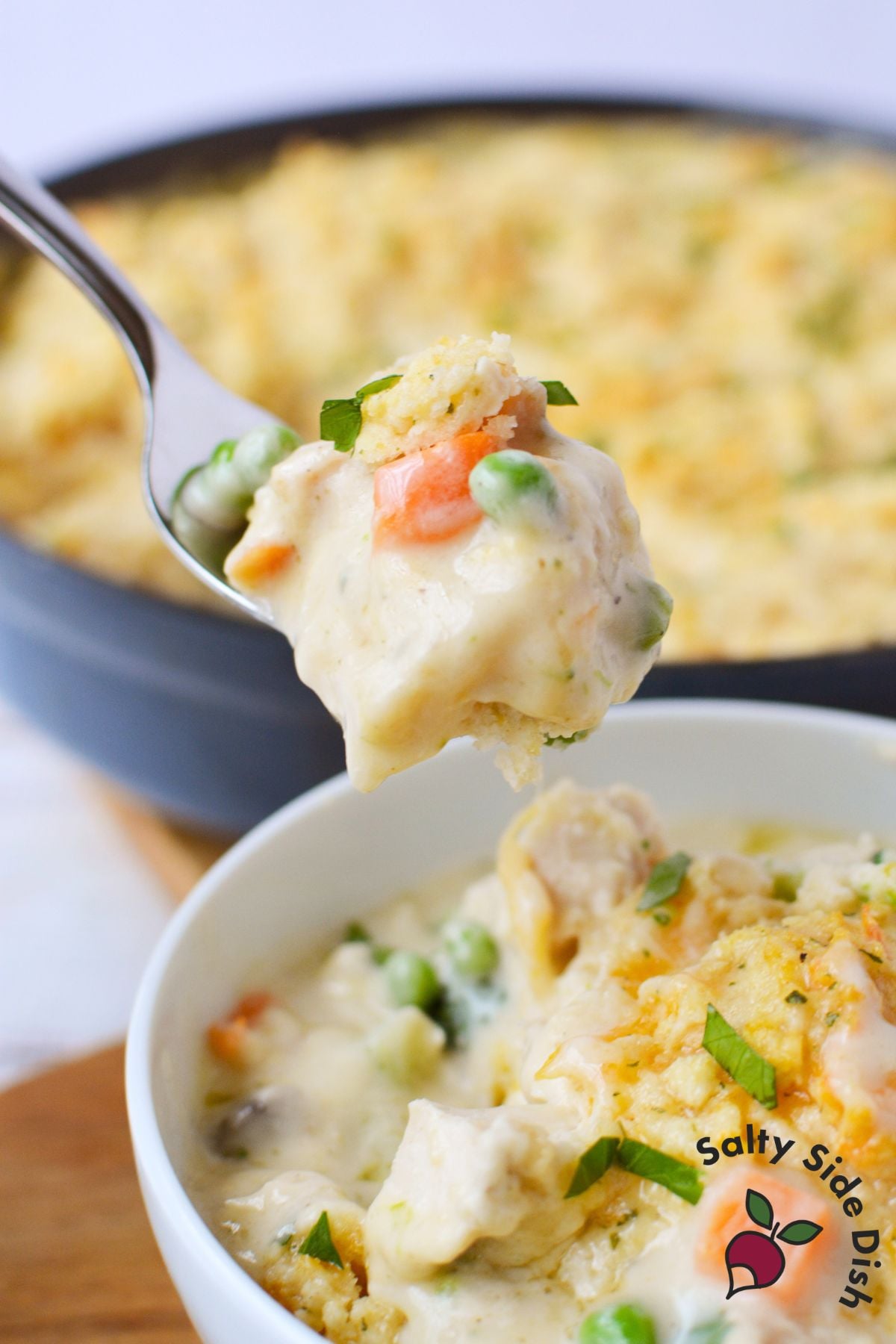 Fork lifting a bite of chicken pot pie with peas, carrots, and biscuit topping over a white bowl filled with a serving of the dish