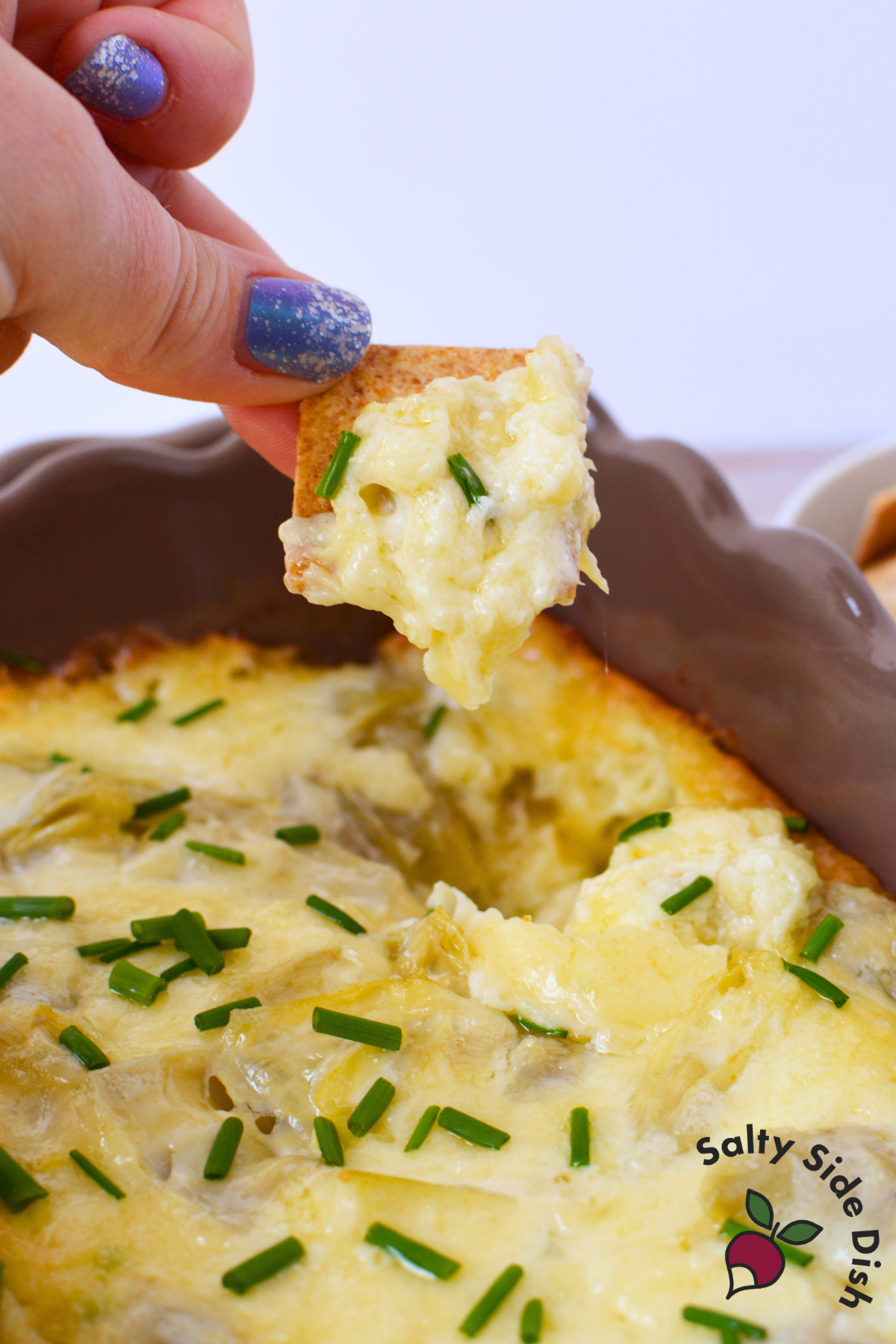cracker being pulled from a round baking dish filled with hot bubbly artichoke dip topped with chives