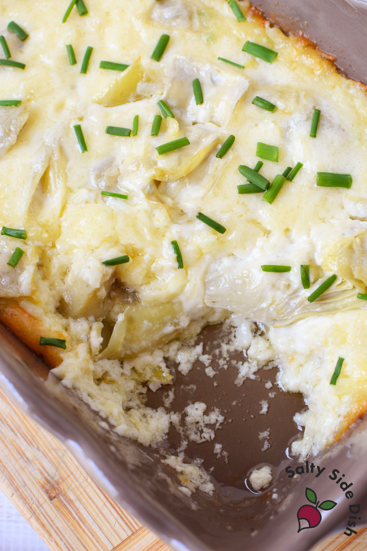 partially scooped baked artichoke dip in a casserole dish showing the creamy cheesy interior with artichoke hearts and chives