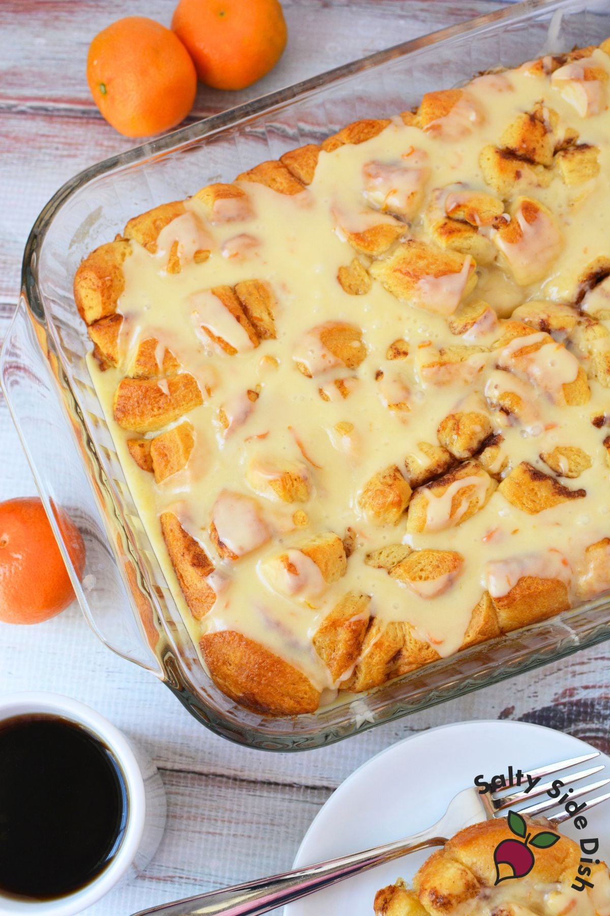 Overhead view of finished orange roll casserole covered in creamy orange glaze in a glass baking dish, surrounded by fresh oranges and a cup of coffee