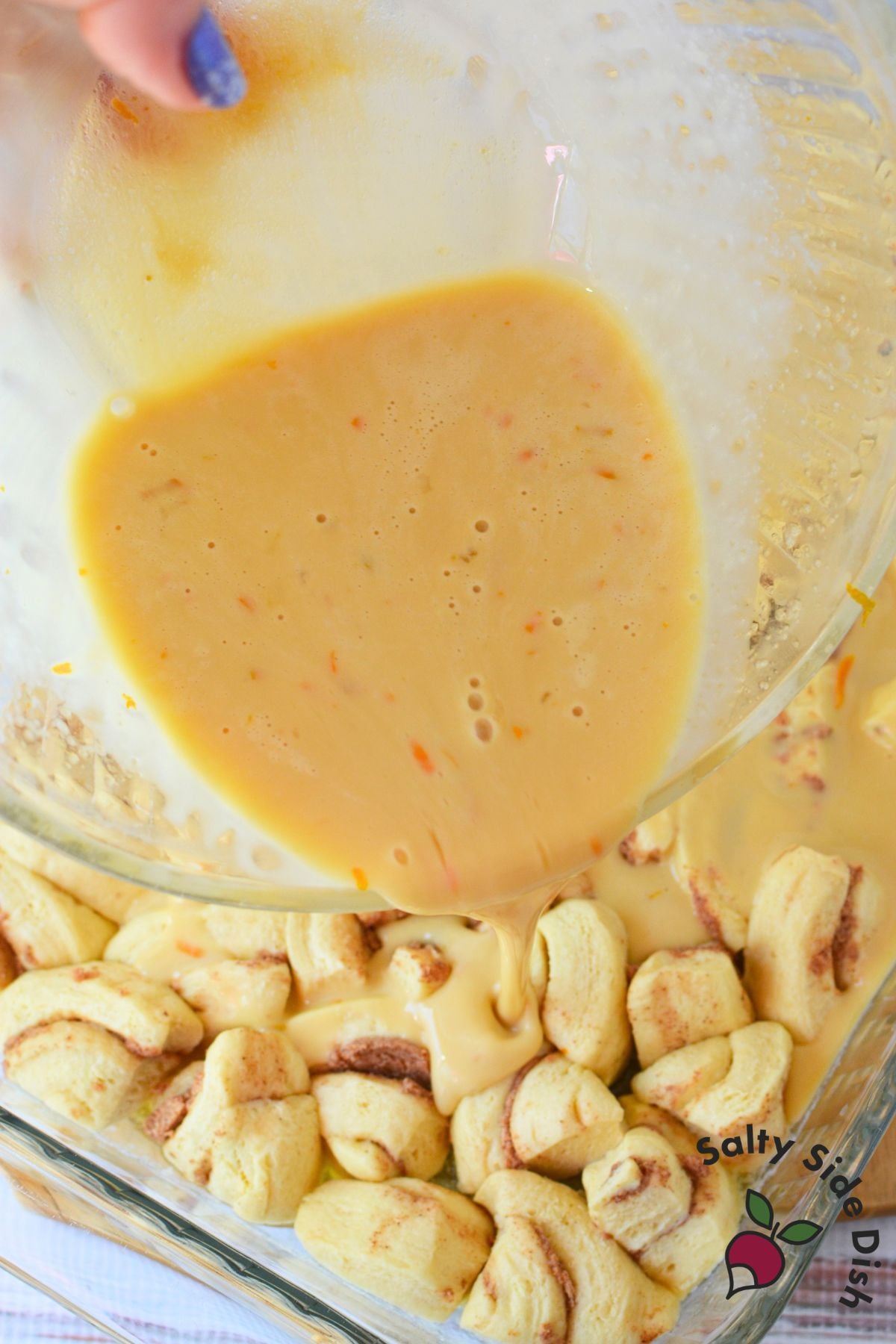 Orange custard mixture being poured from a glass bowl over cut up orange roll pieces in a baking dish