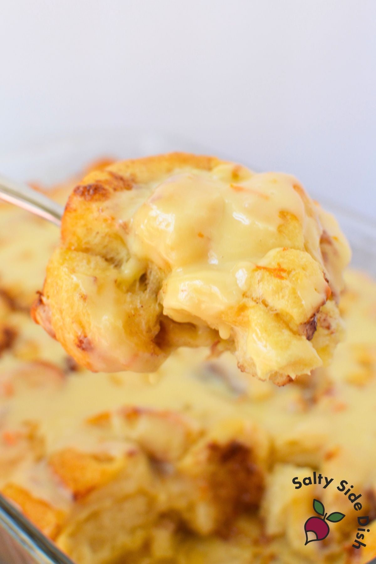 A large spoonful of orange roll casserole dripping with warm vanilla orange glaze held above the baking dish.
