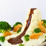 Mashed potato volcano with brown gravy flowing down into a lake surrounded by broccoli and dino nuggets