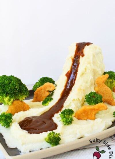 Mashed potato volcano with brown gravy flowing down into a lake surrounded by broccoli and dino nuggets