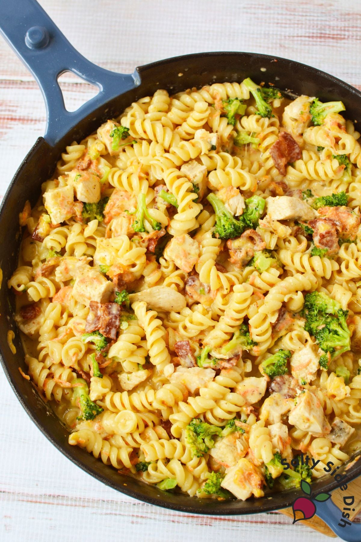 Skillet filled with creamy rotini pasta, chicken, broccoli, bacon, carrots, and melted cheese.