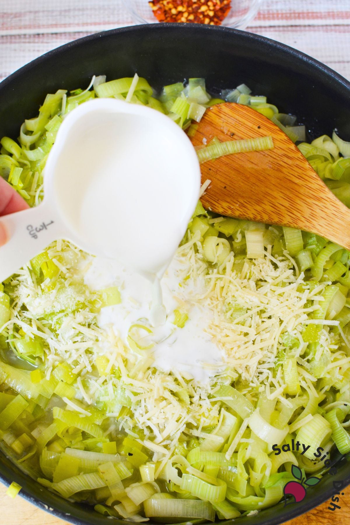 Pouring half and half over cooked leeks topped with shredded parmesan in a skillet