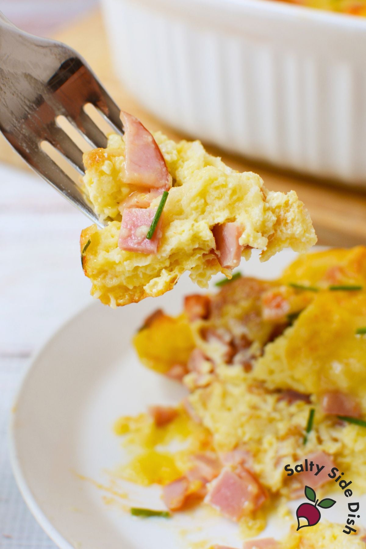 Forkful of eggs benedict casserole with Canadian bacon and chives above the baking dish
